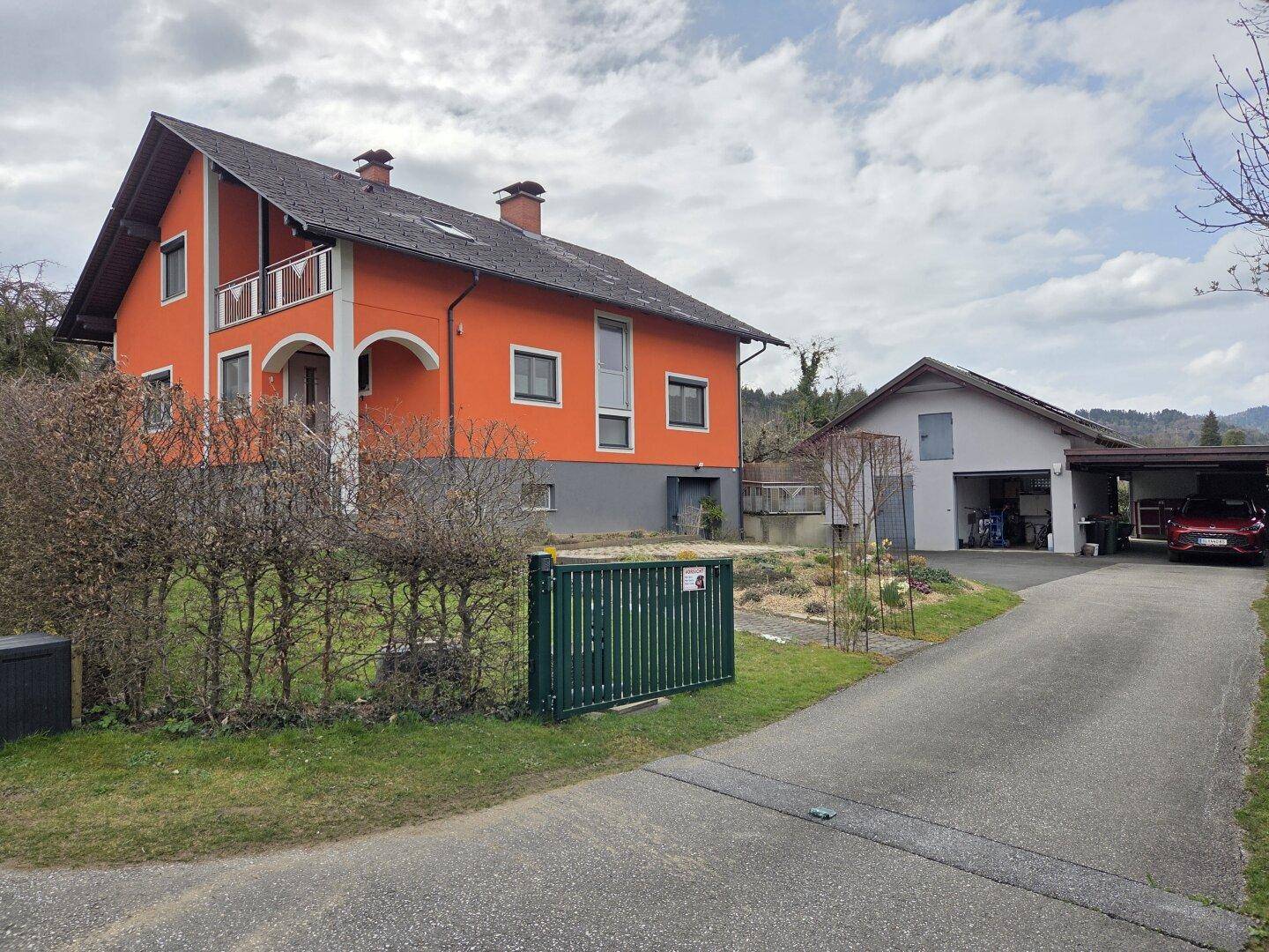 Gesamtansicht des orangefarbenen Hauses mit angrenzender Garage und Carport, umgeben von Grünflächen.