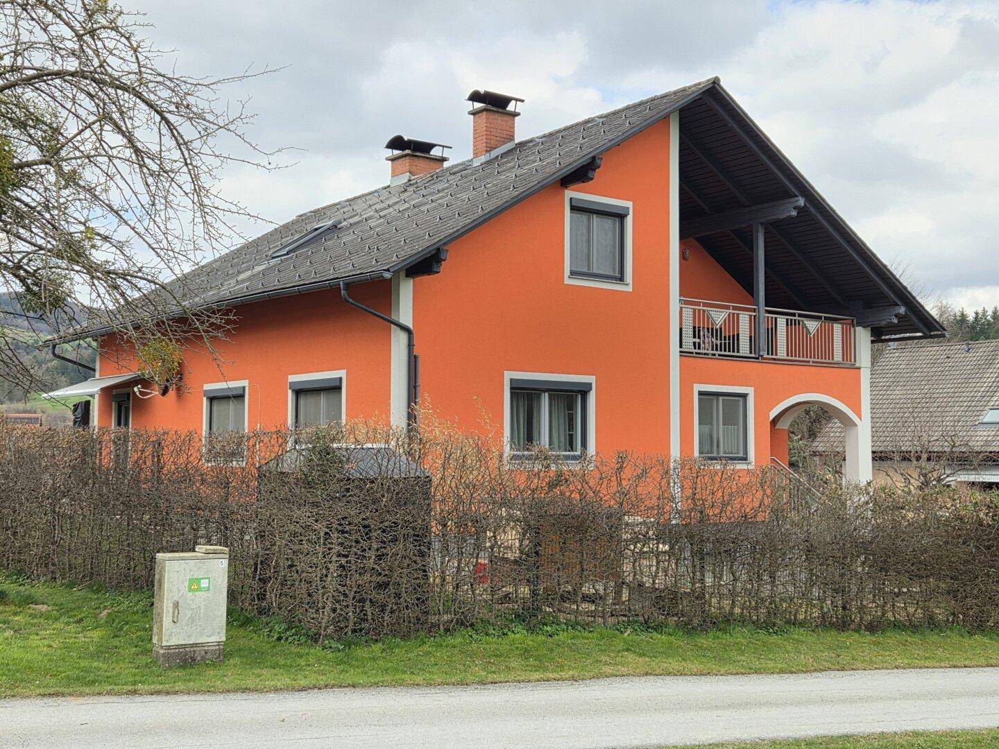 Ansicht des orangefarbenen Einfamilienhauses mit Balkon und Satteldach, umgeben von Hecken und Bäumen.