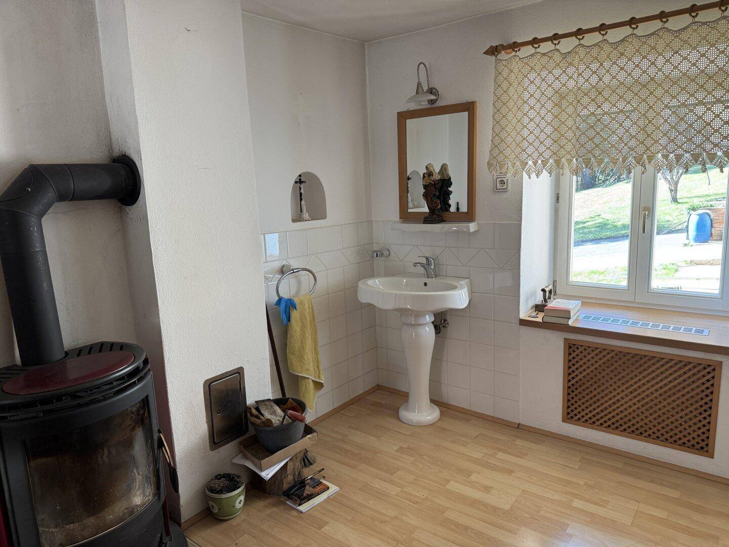 Helles Badezimmer mit Holzofen, Waschbecken und Fenster mit Blick ins Grüne.