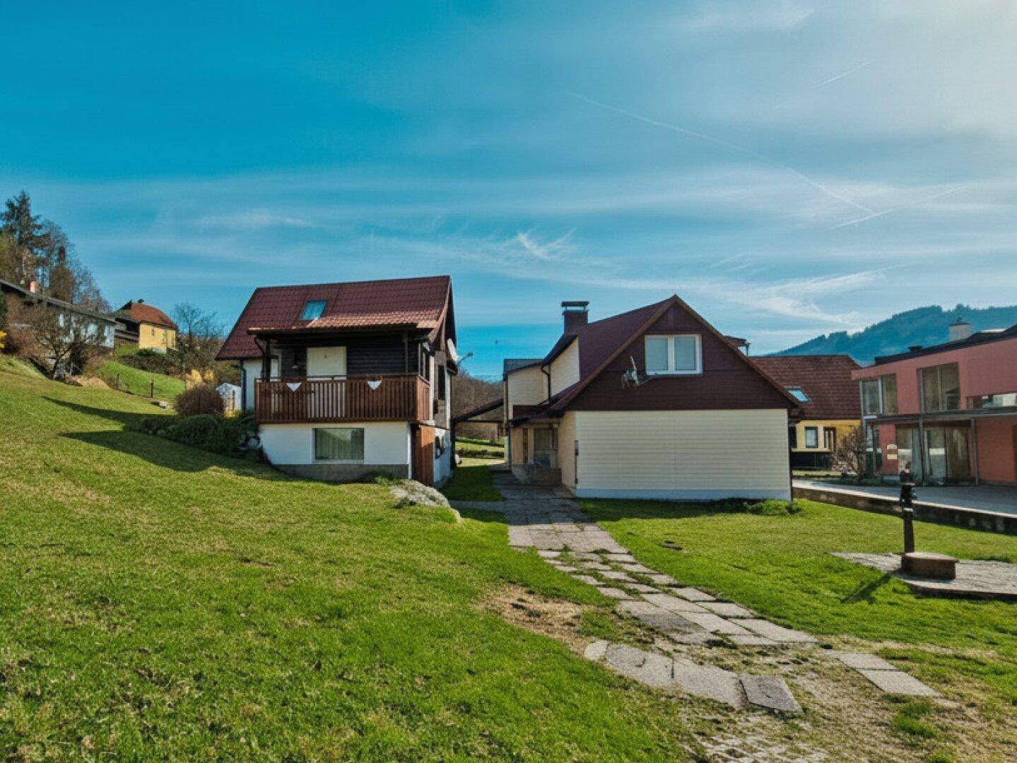 Ansicht von zwei Häusern in einer ländlichen Umgebung mit weitläufigen Grünflächen und blauem Himmel.