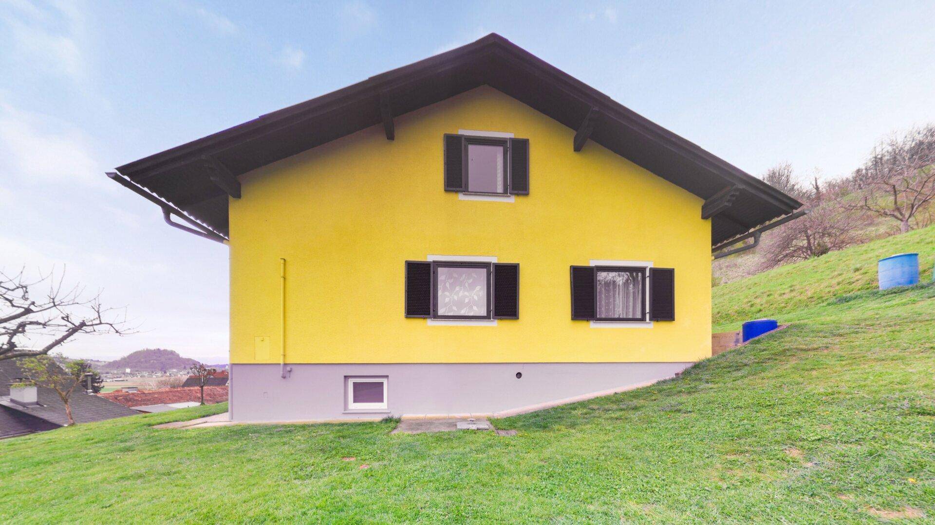 Seitenansicht des gelben Hauses mit schwarzem Dach und grüner Hanglage, umgeben von Natur.