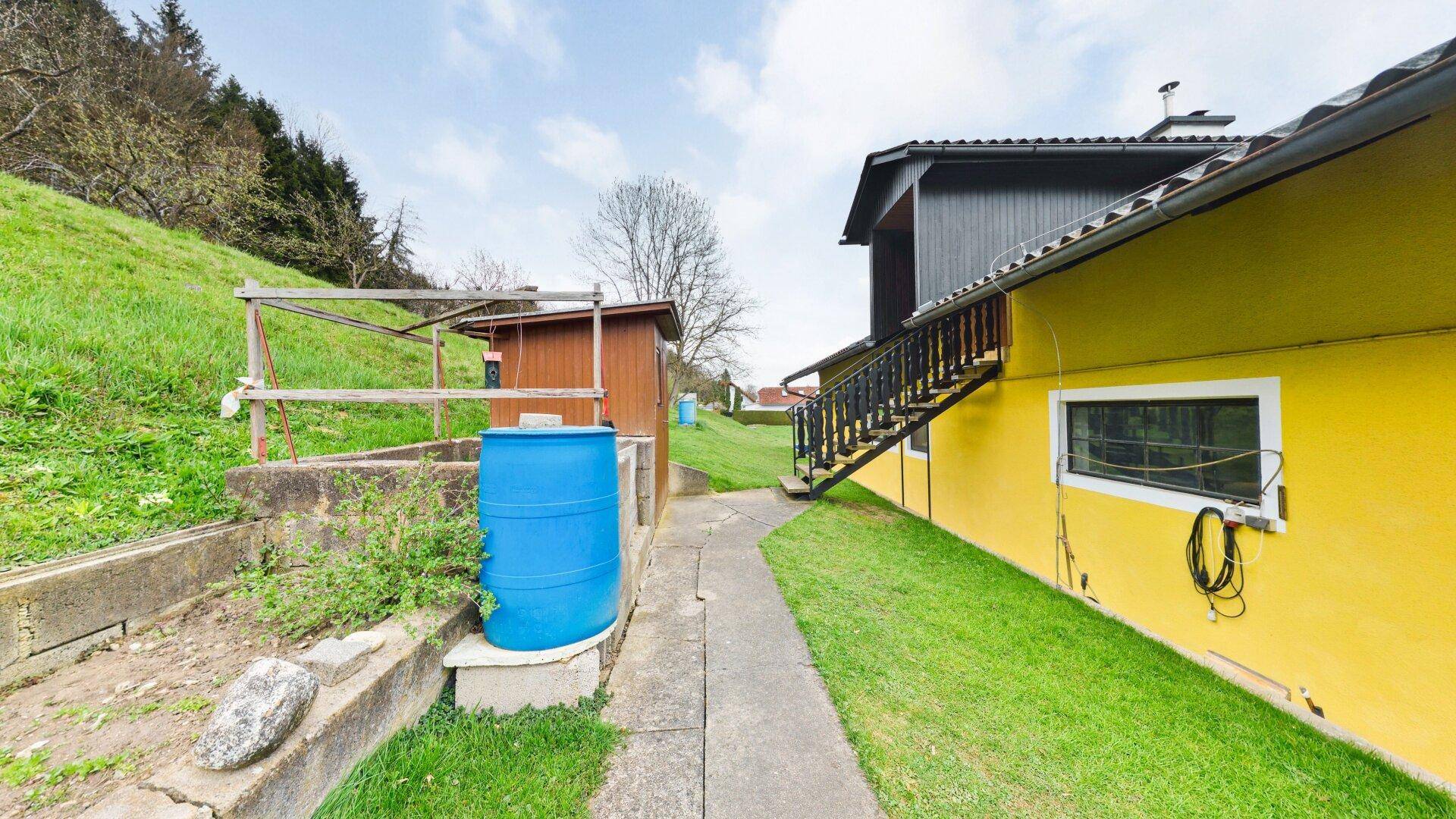 Seitenansicht des Hauses mit Zugang zum Garten und einem blauen Regenfass auf dem Grundstück.