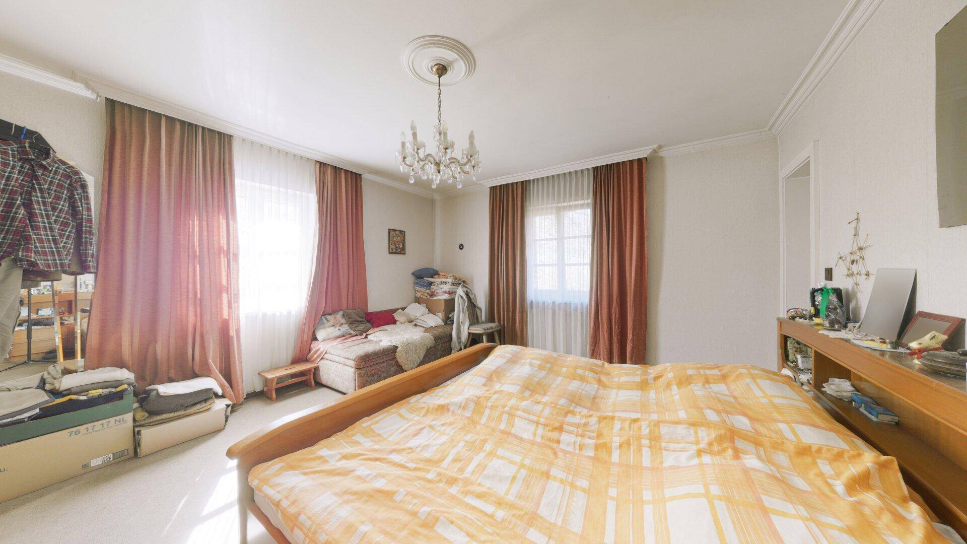 Helles Schlafzimmer mit Doppelbett, Parkettboden und Kronleuchter, gut beleuchtet durch zwei Fenster.