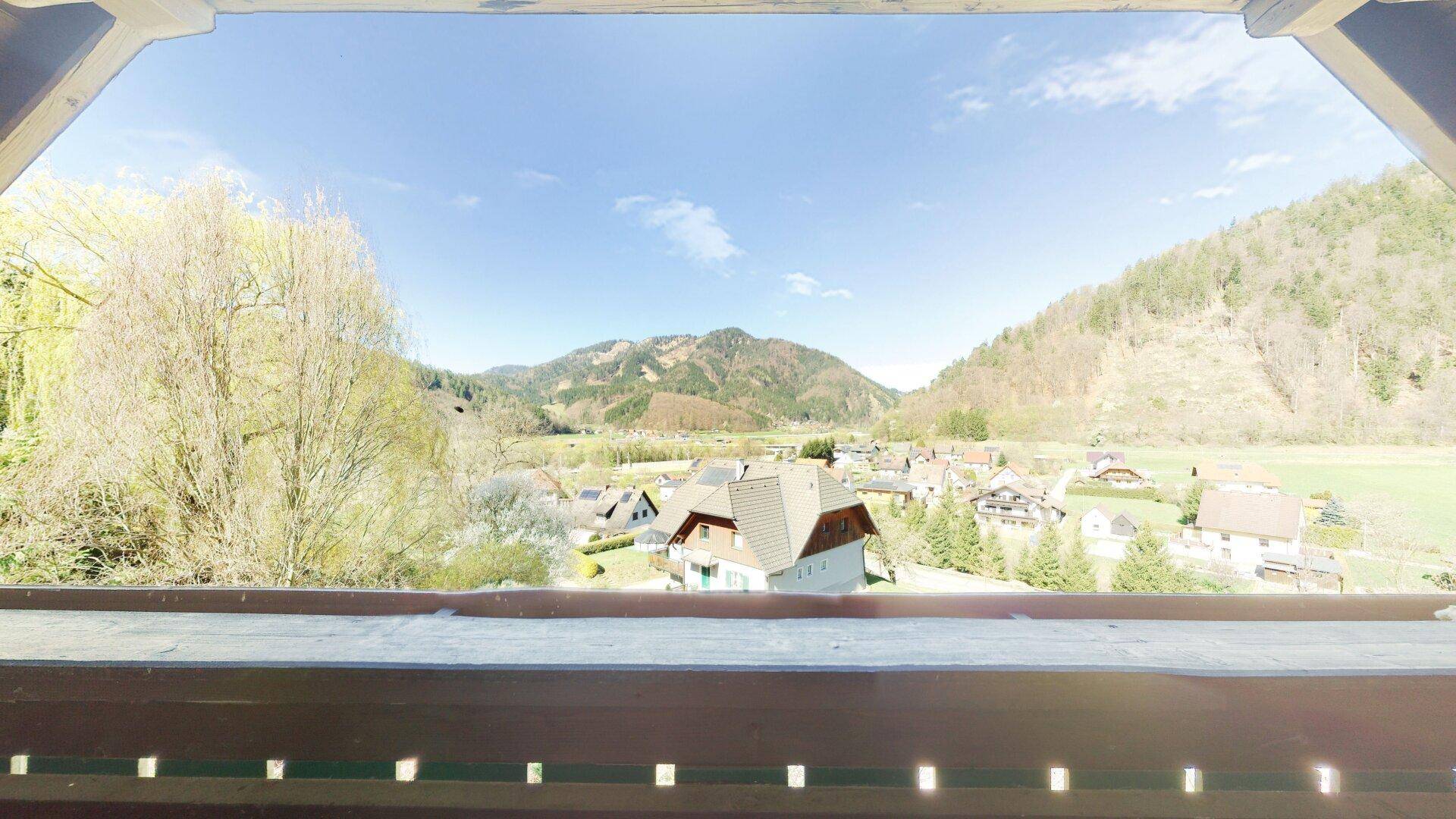 Weitblick von einem Balkon über ein Dorf und die umliegende bergige Landschaft unter blauem Himmel.