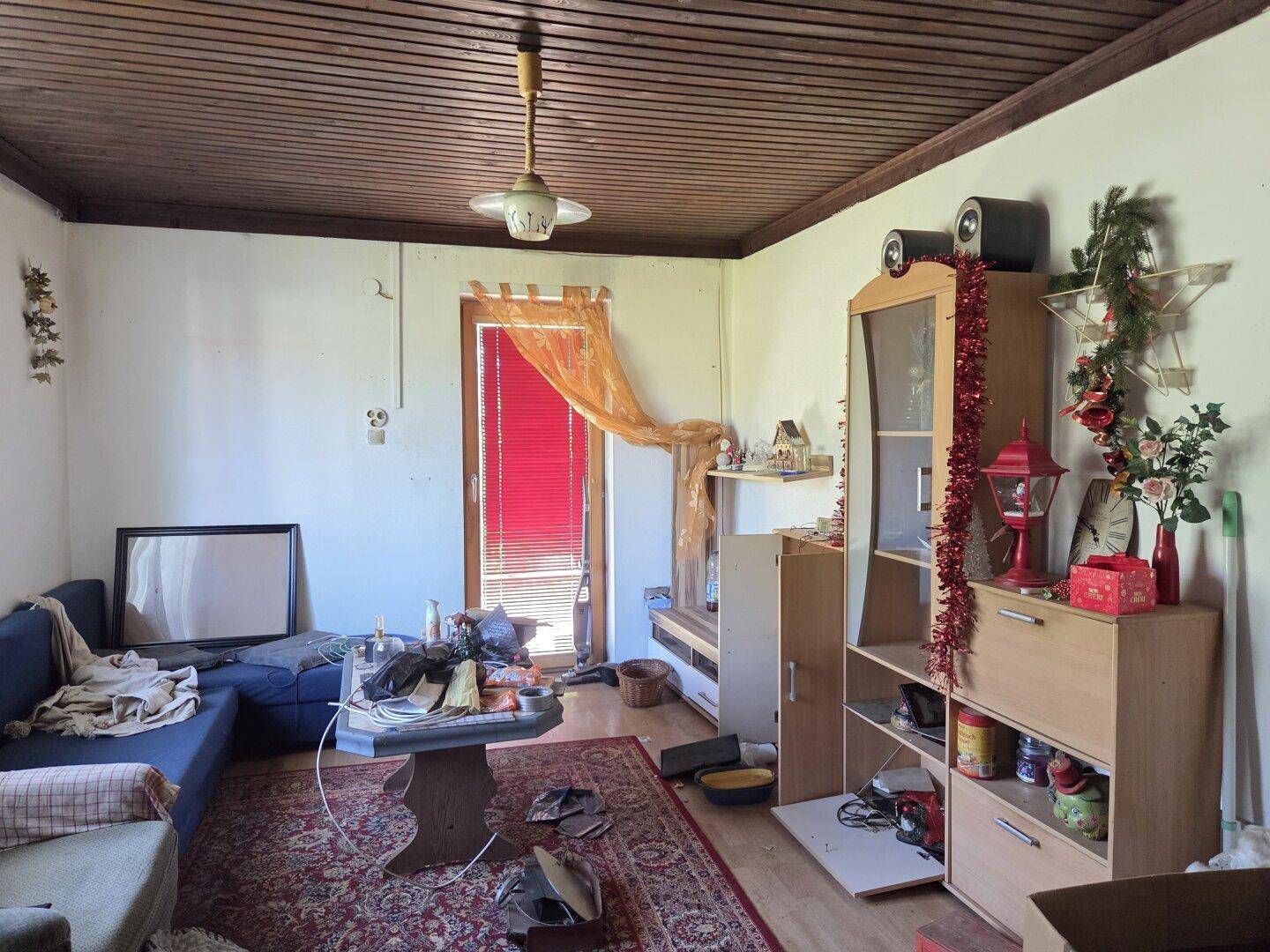 Unaufgeräumtes Wohnzimmer mit Holzdecke, Teppichboden und Blick ins Grüne durch die Tür.