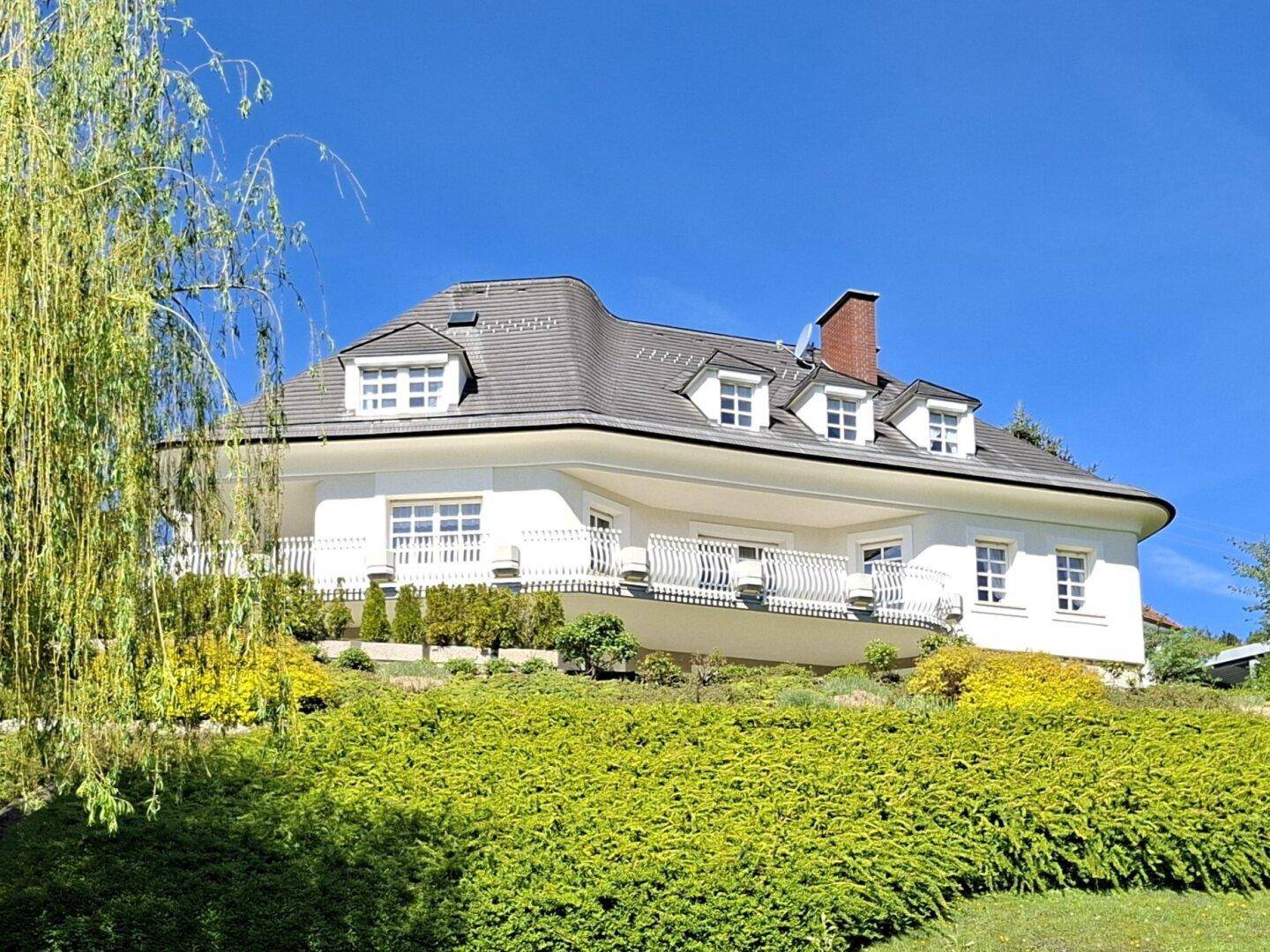 Herrschaftliche Villa mit geschwungenen Balkonen und üppiger grüner Bepflanzung in sonniger Lage.