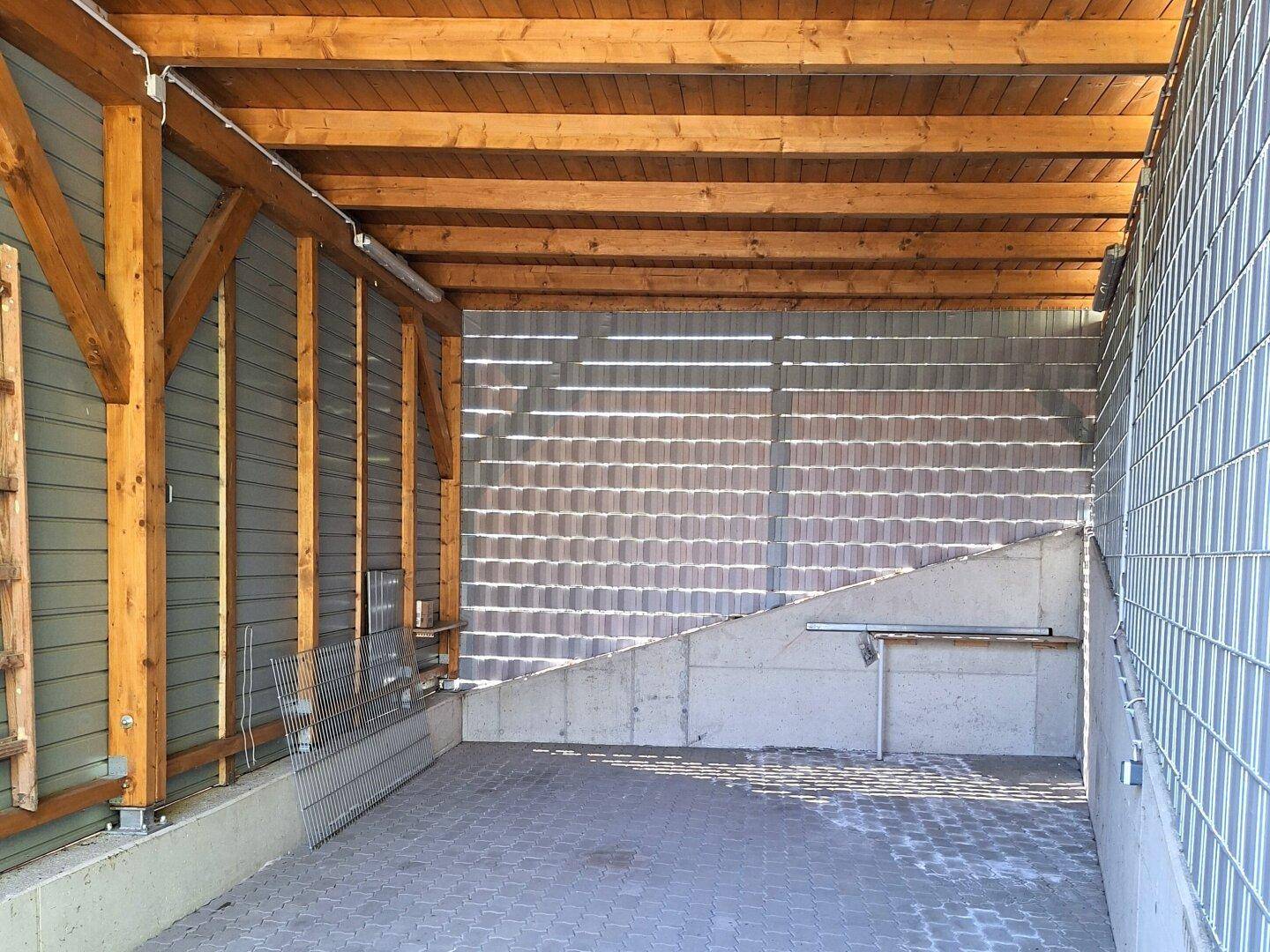 Überdachter Carport mit Holzbalken und gepflastertem Boden, bietet Schutz für Fahrzeuge.