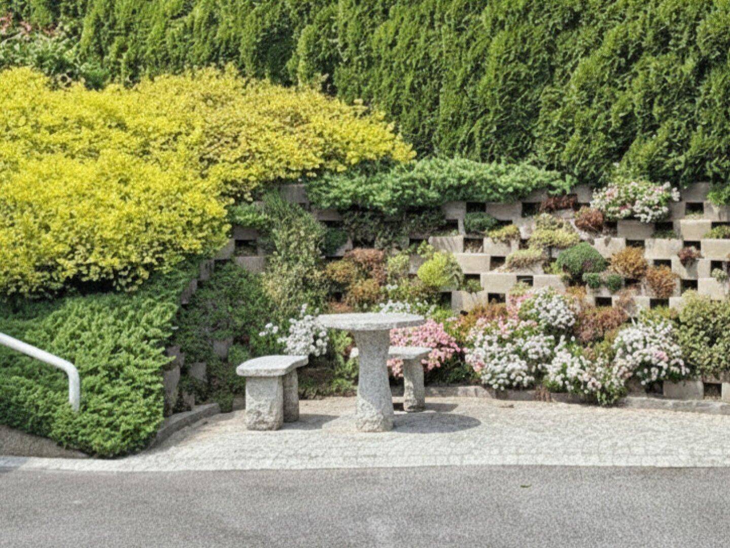 Gepflegter Gartenbereich mit Steinmauer, üppiger Bepflanzung und kleiner Sitzgelegenheit aus Stein.