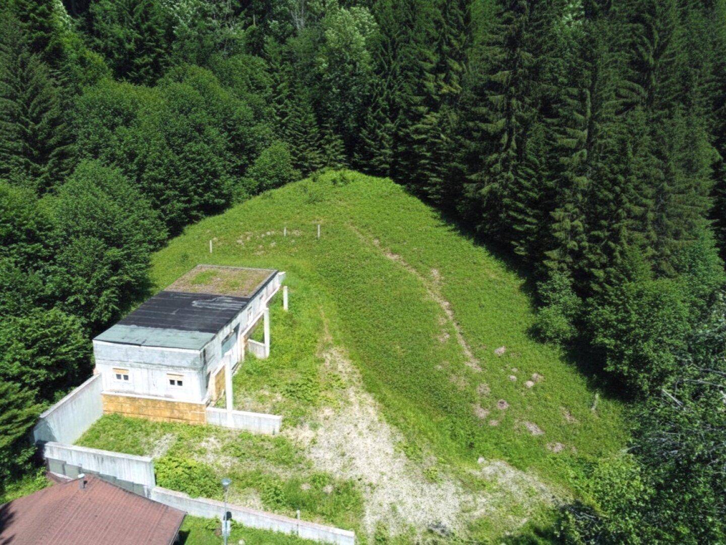 Luftaufnahme des Gebäudes mit Flachdach, umgeben von dichtem Wald und einer grünen Hügellandschaft.