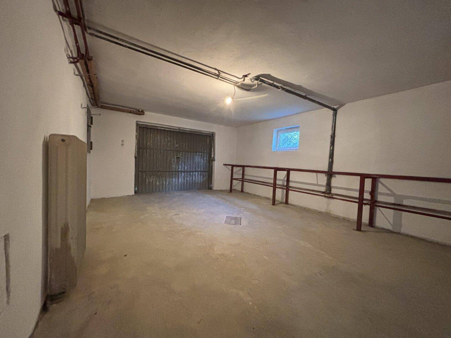 Geräumige Garage im Untergeschoss mit Metalltoren und sichtbaren Leitungen.