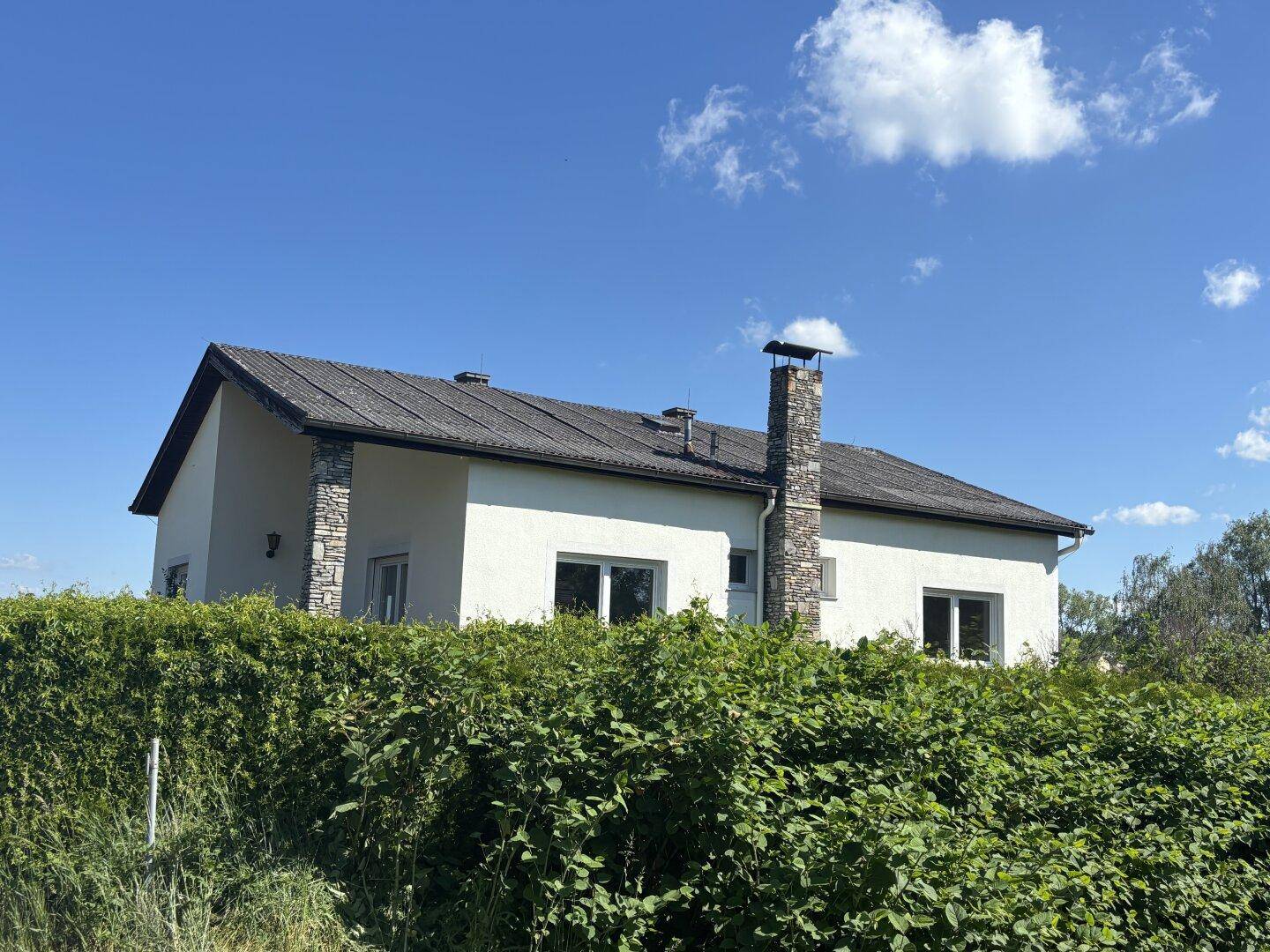 Seitenansicht des Hauses mit Schornstein und grüner Umgebung unter blauem Himmel.