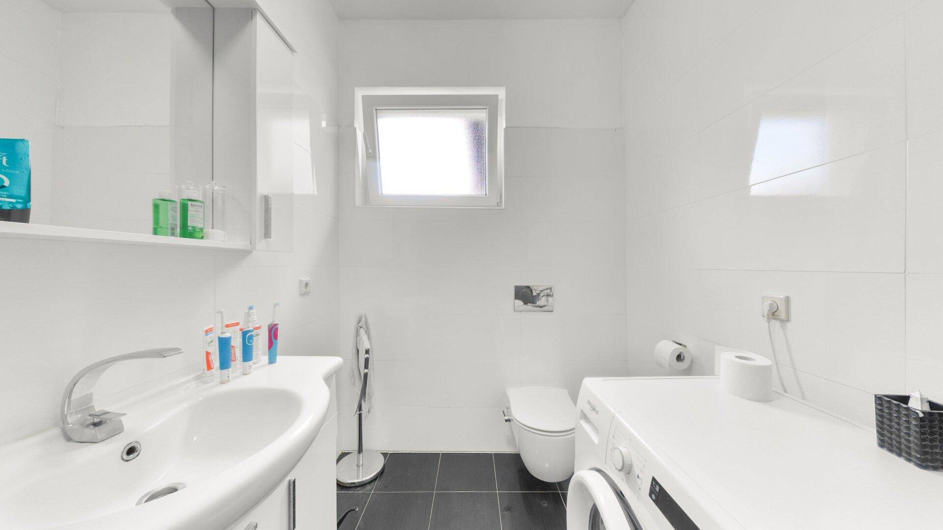 Modernes Badezimmer mit Waschbecken, Toilette, Waschmaschine und Fenster, hell gefliest.