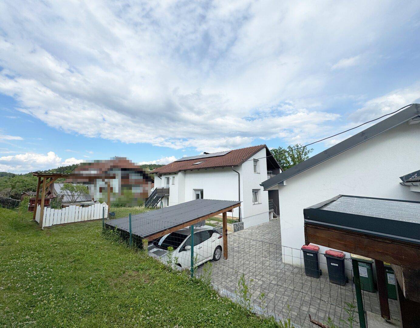 Haus mit Carport und Gartenbereich, umgeben von grüner Landschaft unter blauem Himmel.