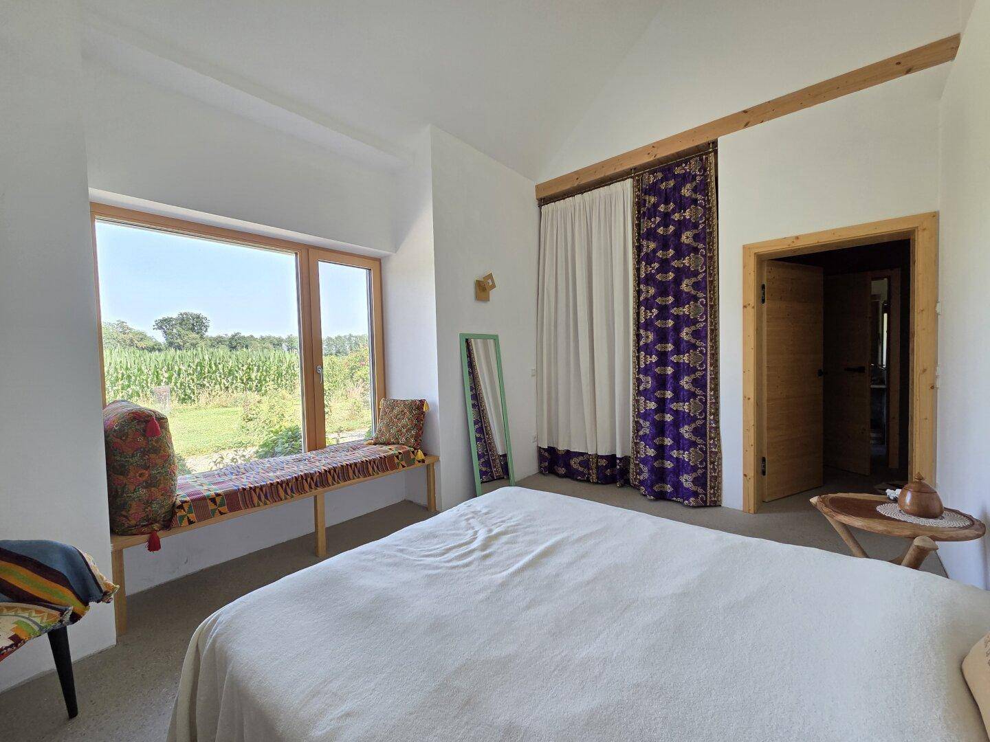 Helles Schlafzimmer mit großer Fensterfront, Sitzbank und Blick ins Grüne.