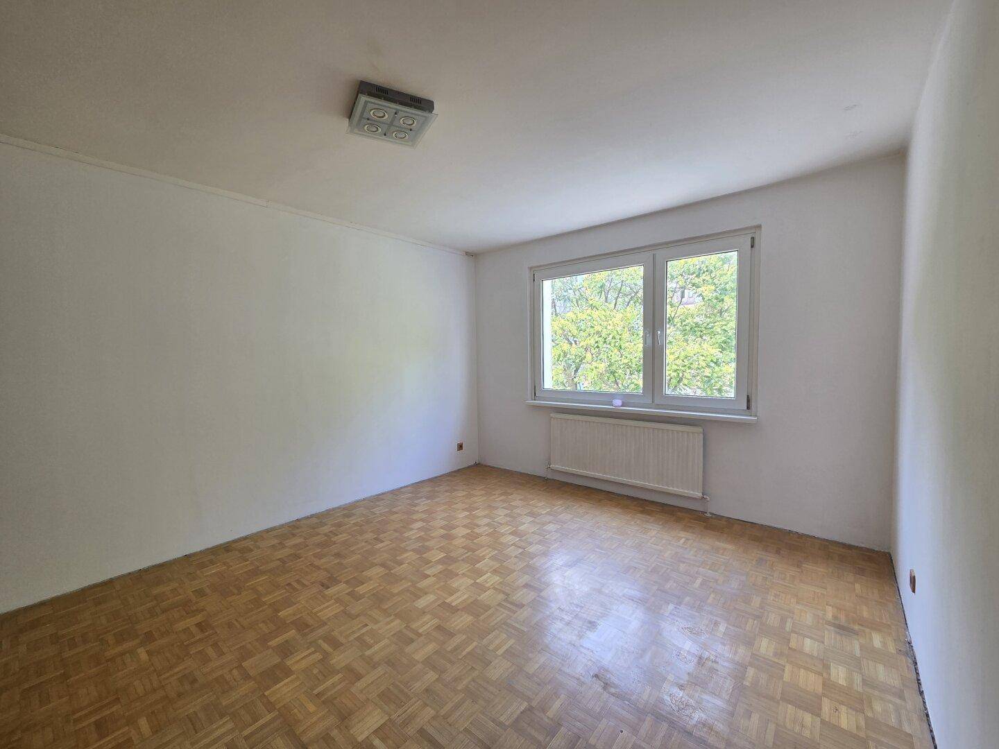 Helles Schlafzimmer mit Parkettboden und großem Fenster mit Blick ins Grüne.