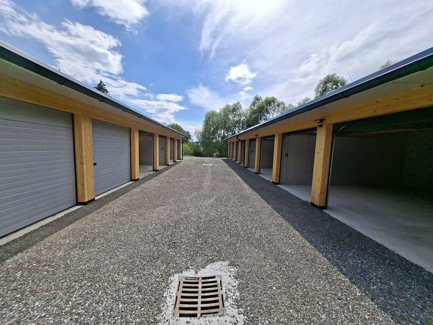 Blick auf eine Reihe von Garagenboxen mit grauen Toren und Holzelementen entlang eines Schotterwegs.