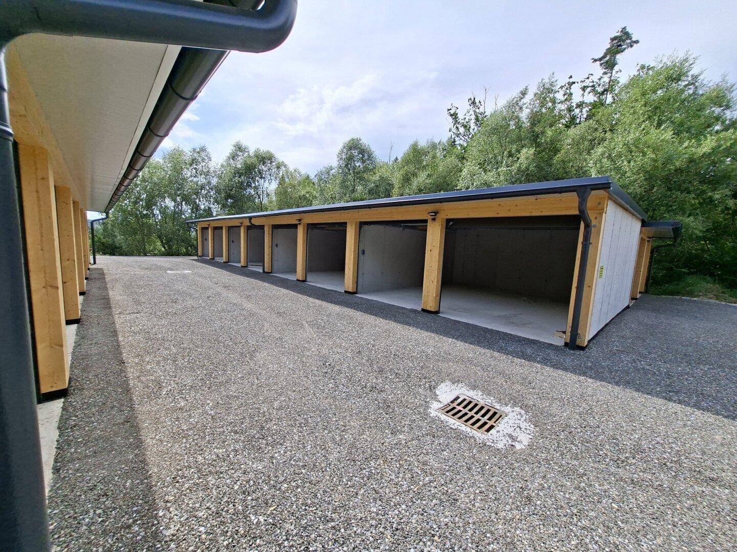 Seitenansicht einer Reihe offener Garagenboxen mit Betonwänden und Holzbalken, davor ein Schotterweg.