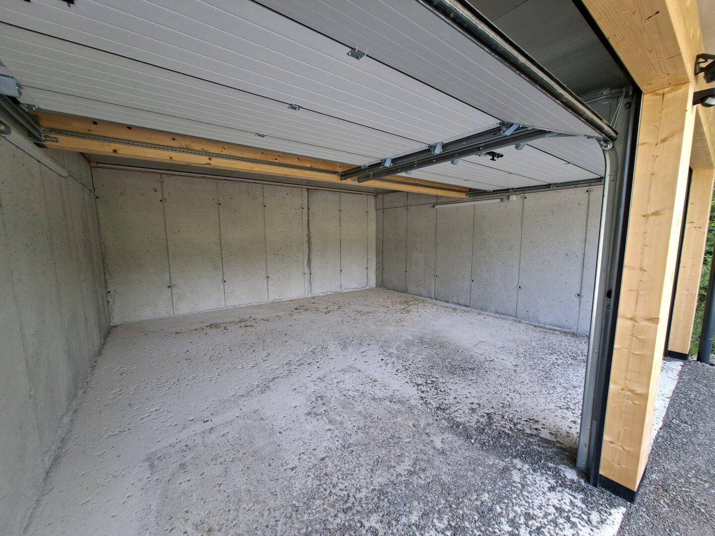 Innenansicht einer leeren Garagenbox mit Betonwänden und -boden sowie einem Sektionaltor.