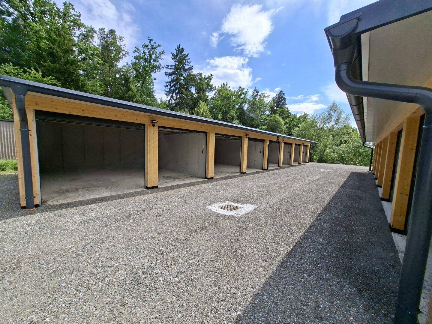 Reihe von Garagenboxen mit offenen Eingängen, Betonwänden und sichtbaren Holzstützen unter blauem Himmel.