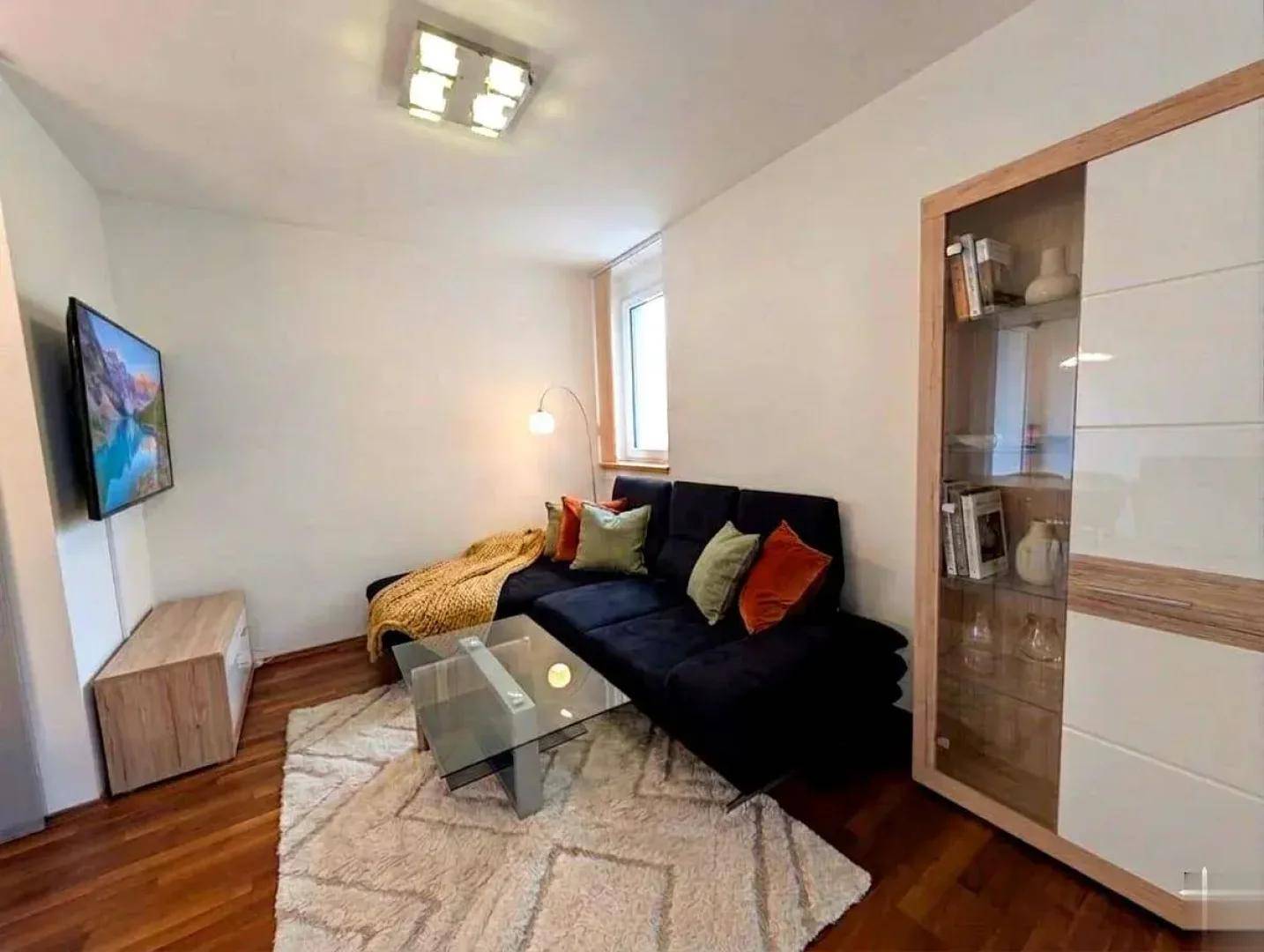 Gemütliches Wohnzimmer mit dunklem Ecksofa, Fernseher und hellem Teppich.