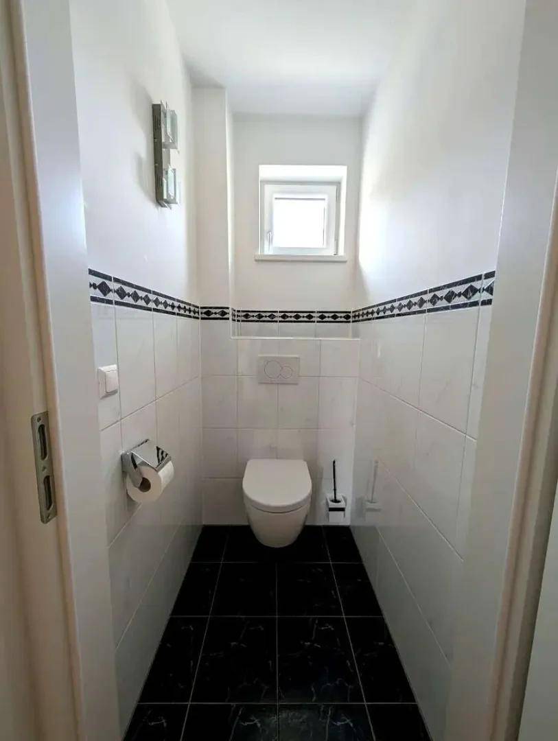 Kleines Gäste-WC mit Fenster, Wand-WC und dunklen Bodenfliesen.