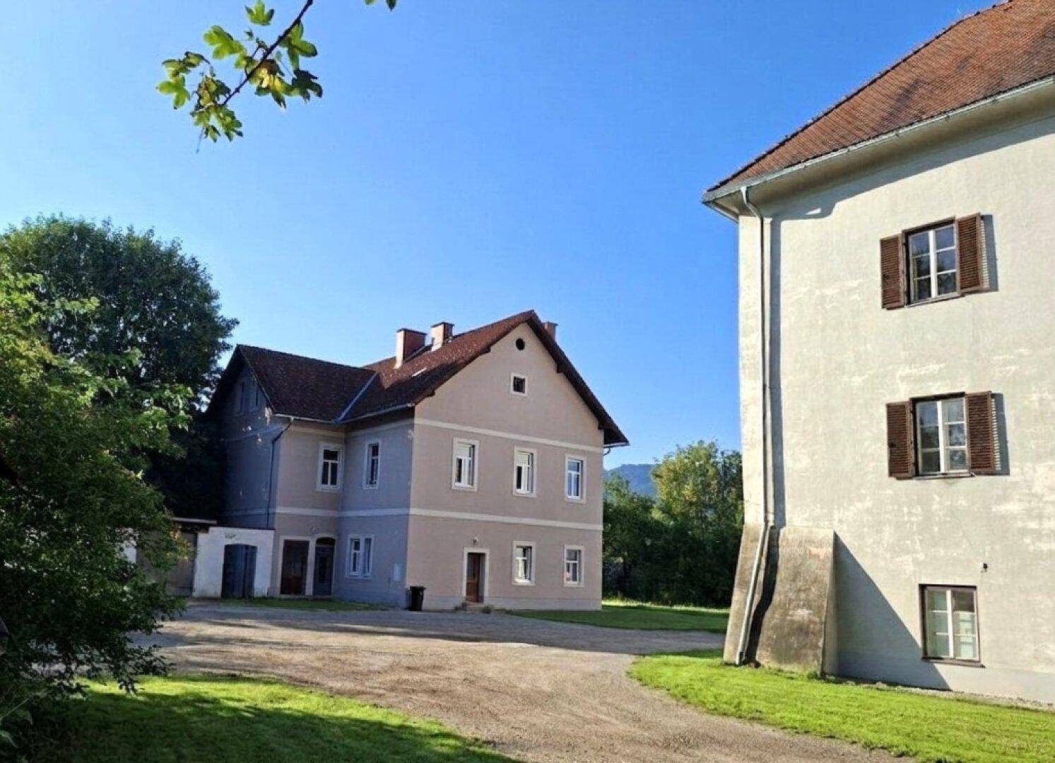 Ansicht von zwei historischen Gebäuden auf einem weitläufigen Grundstück mit Schotterfläche und Grün.