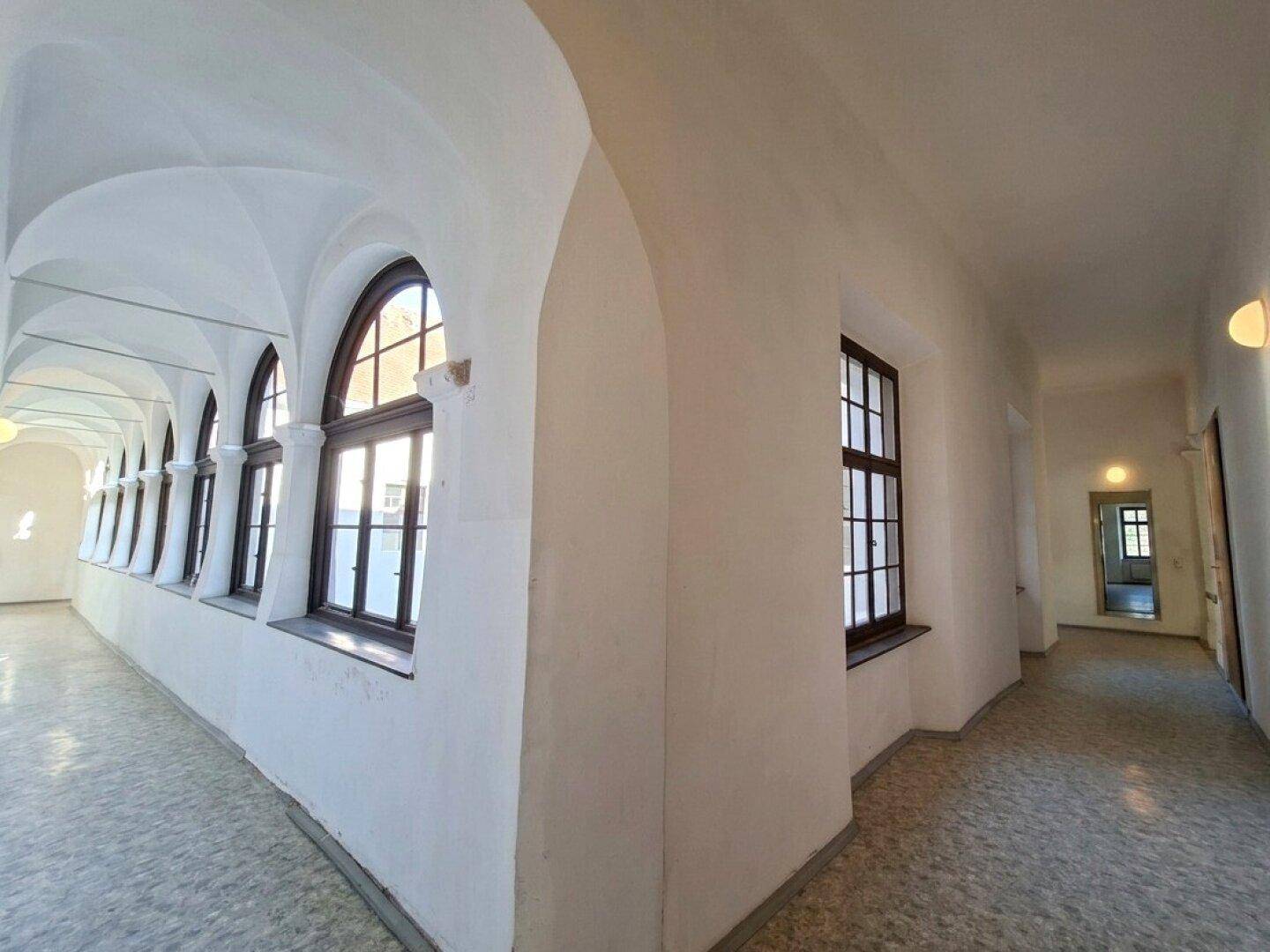 Langer Flur mit Gewölbedecken, hohen Fenstern und hellem Steinboden, der viel Licht hereinlässt.