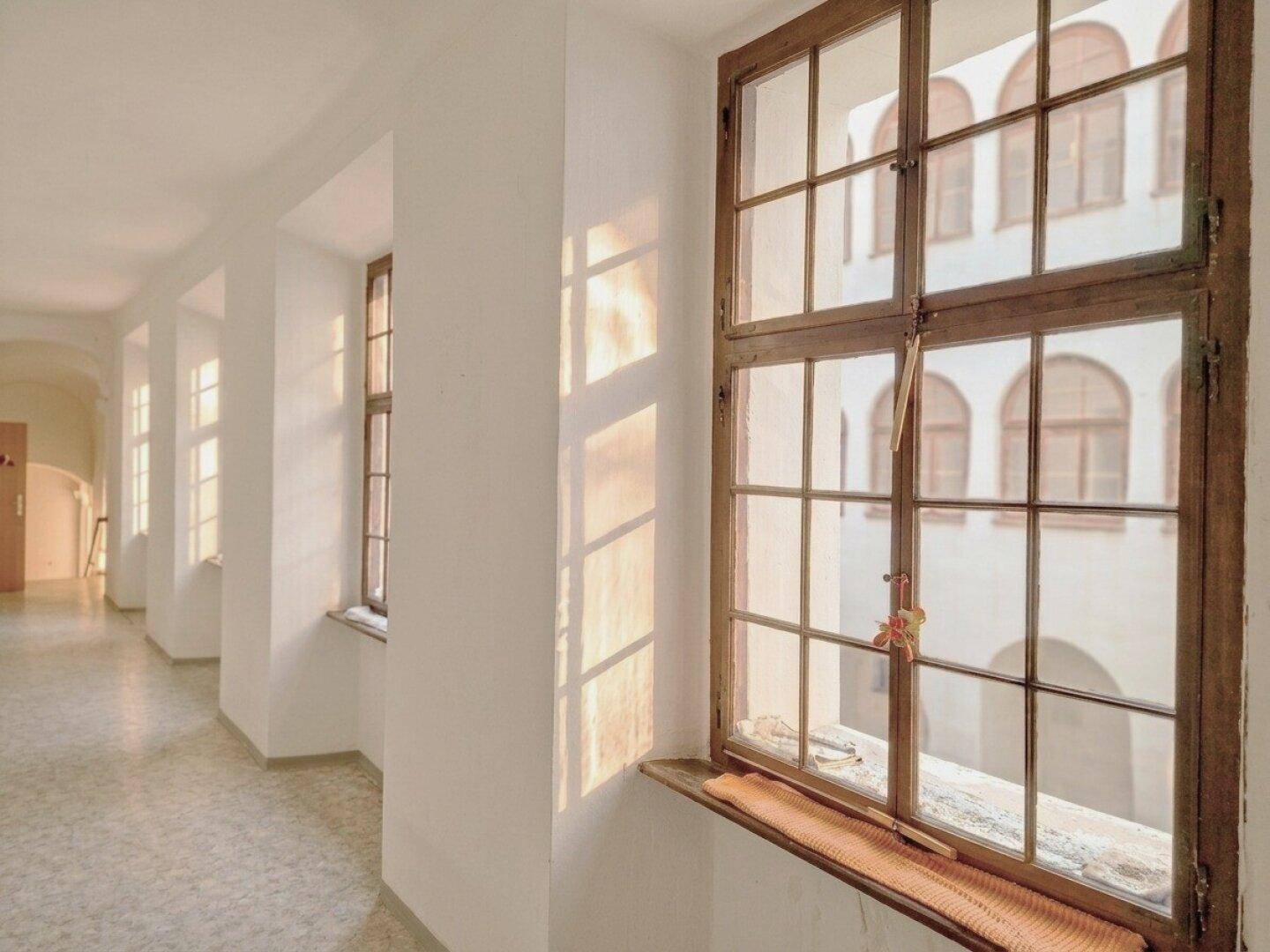 Sonnenlicht durchflutet den Flur durch ein großes Holzfenster mit Blick auf den Innenhof.