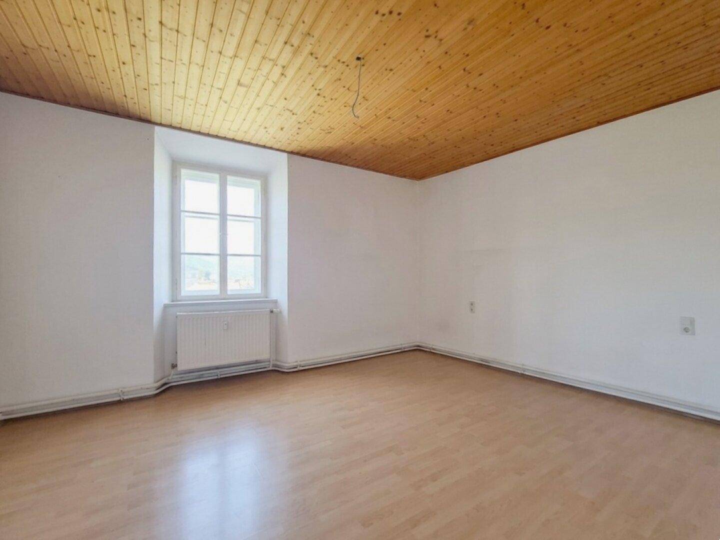 Heller Raum mit Holzdecken und -böden, einem Fenster und einem Heizkörper darunter.