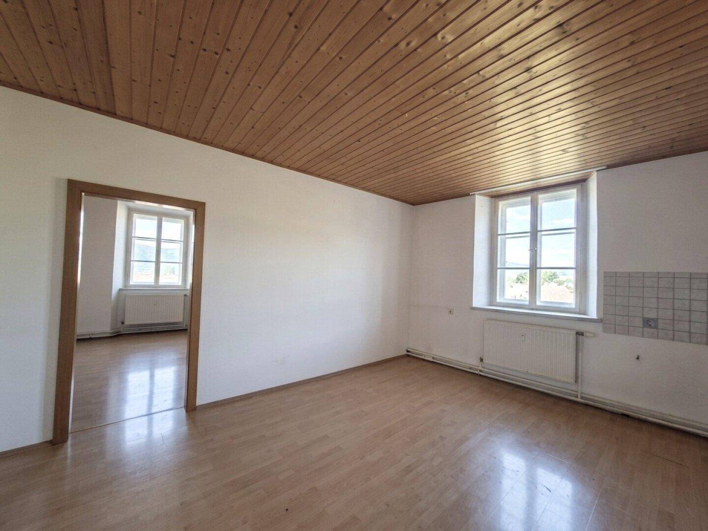 Heller Raum mit Holzdecken und -böden, einem Fenster und Zugang zu einem weiteren Zimmer.