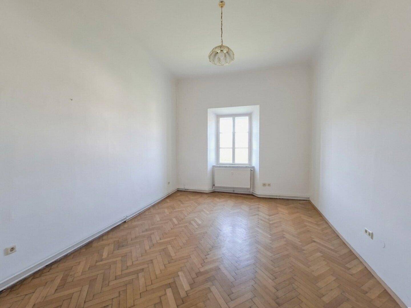 Leerer Raum mit weiß gestrichenen Wänden, Fischgrätparkett und einem Fenster mit Heizkörper.