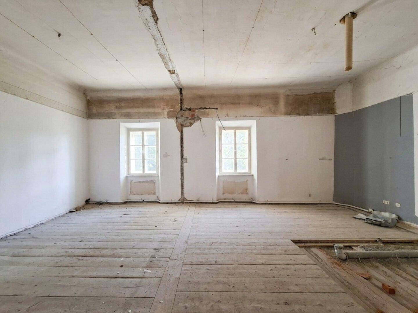 Renovierungsbedürftiger Raum mit Holzdielenboden, beschädigten Wänden und zwei Fenstern mit Heizkörpern.