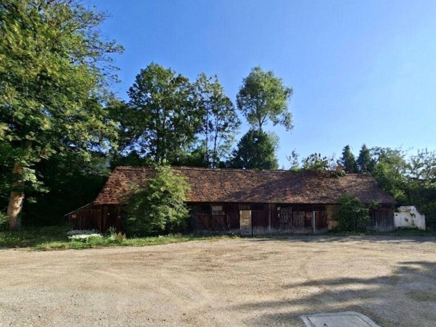 Altes Scheunengebäude mit braunem Ziegeldach und Holzfassade, umgeben von Bäumen und Schotter.