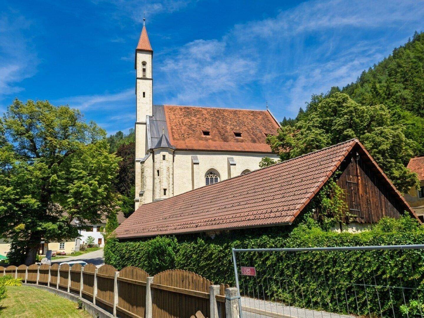 Historische Kirche mit spitzem Turm und umliegender grüner Landschaft unter blauem Himmel.