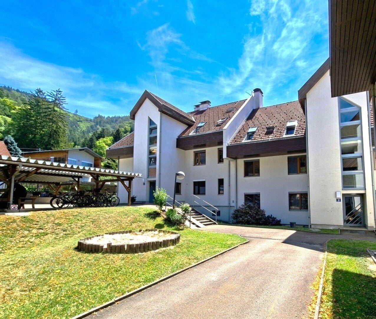 Mehrfamilienhaus mit Carport und Rasenfläche, umgeben von grünen Hügeln unter strahlend blauem Himmel.