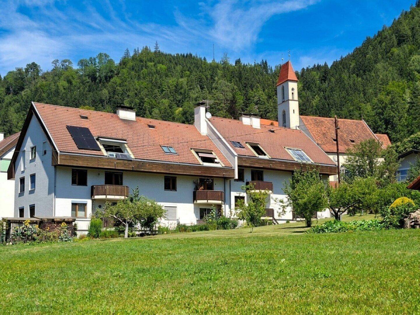 Wohngebäude mit Balkonen und Solaranlagen auf dem Dach, umgeben von grünen Wiesen und Bergen.