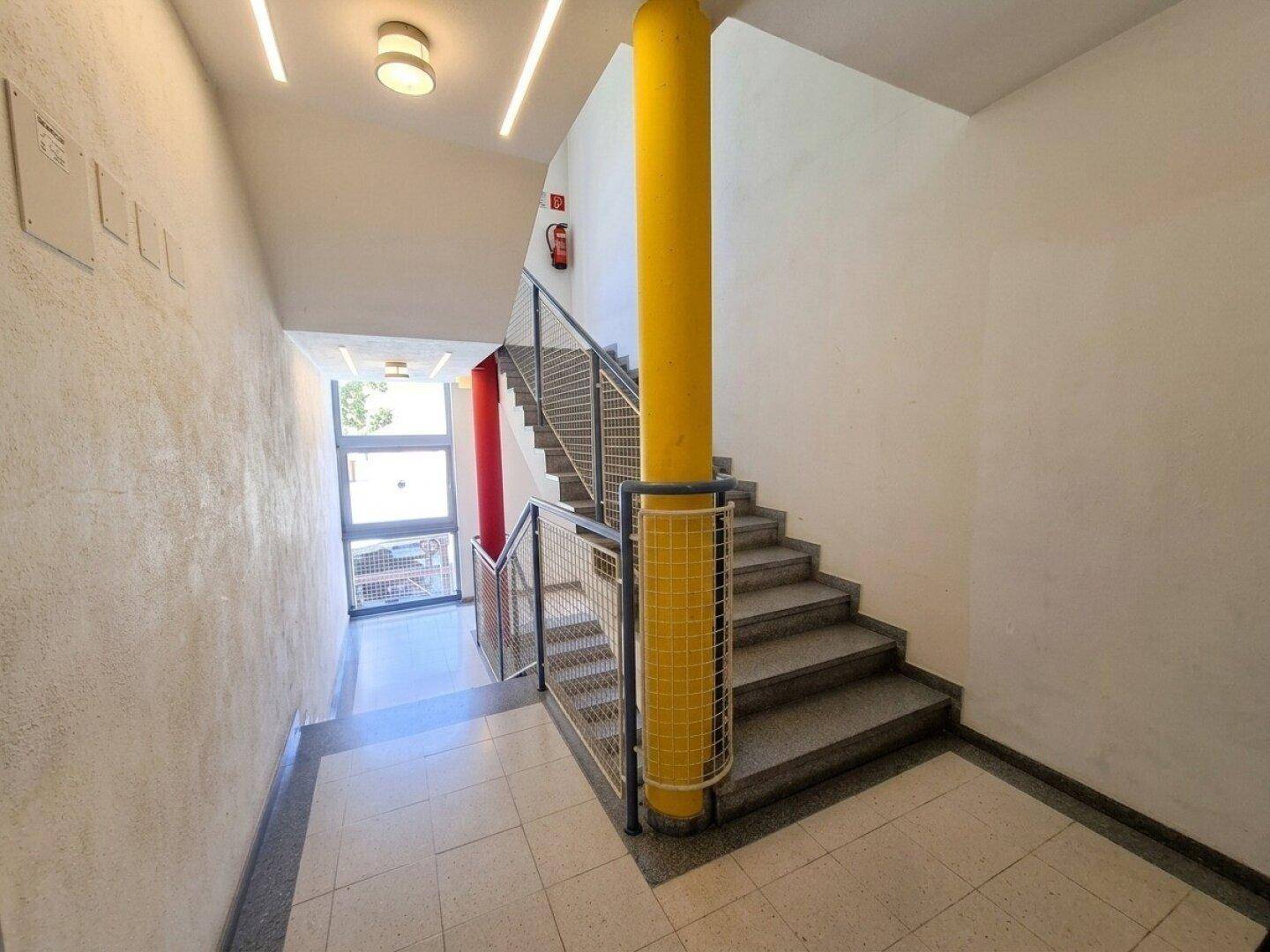 Heller Treppenaufgang mit gelbem Pfeiler, Metallgeländer und modernen Leuchten im Eingangsbereich.