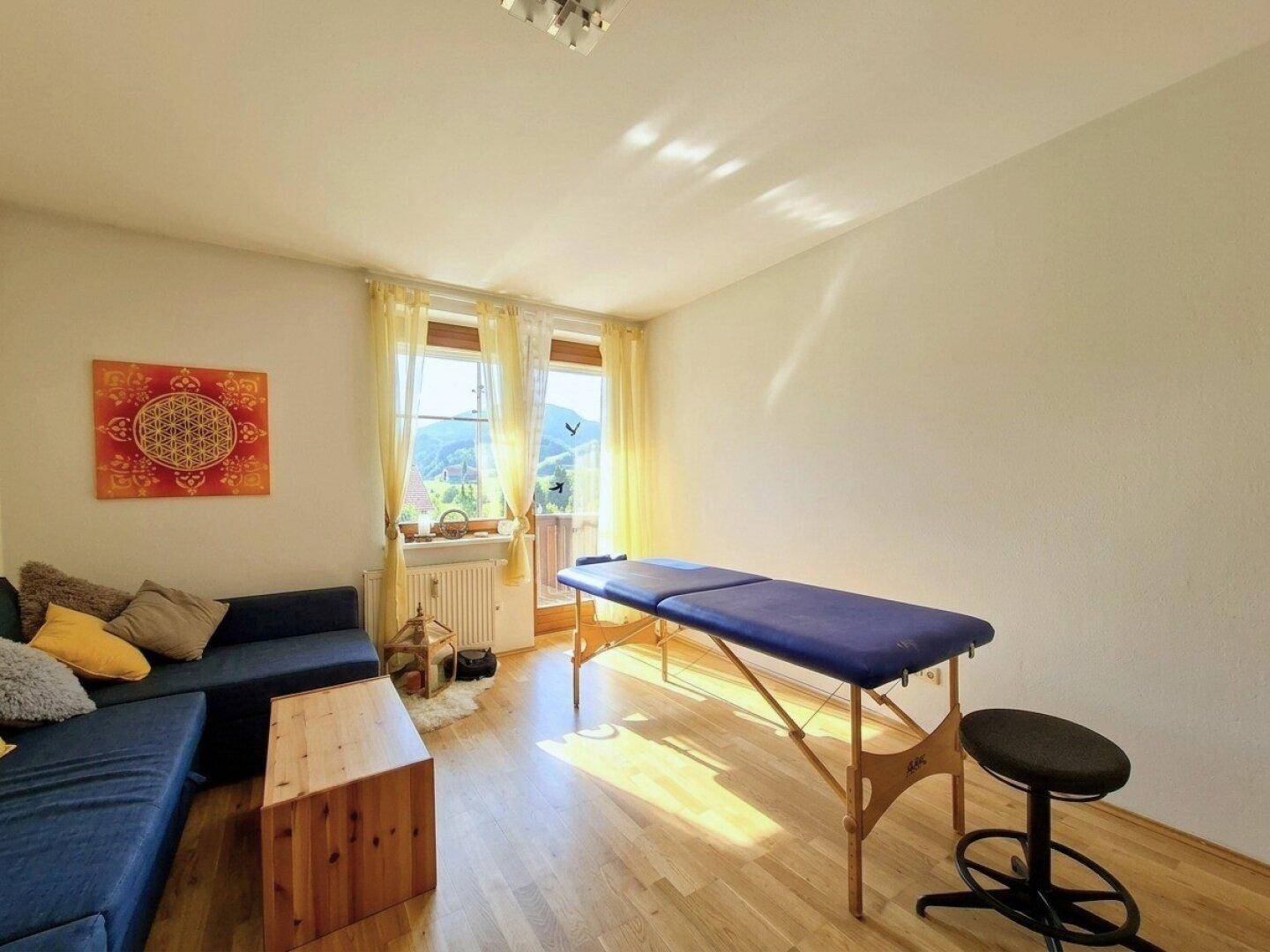 Heller Raum mit Sofa, Massageliege und Fenster, das einen schönen Blick auf die grüne Landschaft bietet.