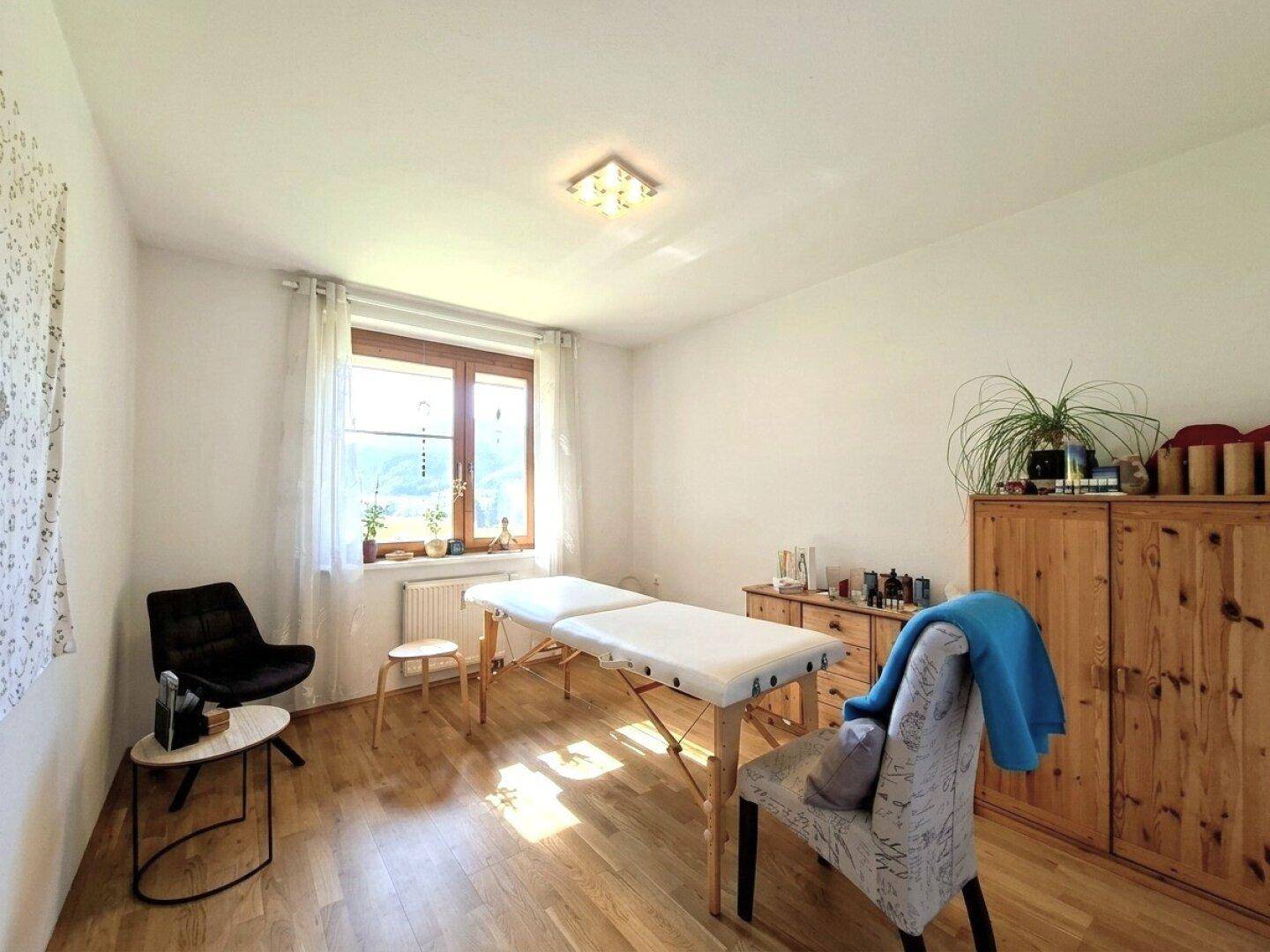 Heller Raum mit Massageliege, Holzmöbeln und Fenster, das viel Tageslicht hereinlässt und eine grüne Aussicht bietet.