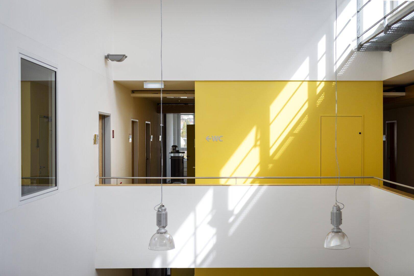 Heller Flur und Treppenhaus mit gelber Akzentwand und modernen Hängelampen in einem Bürogebäude.