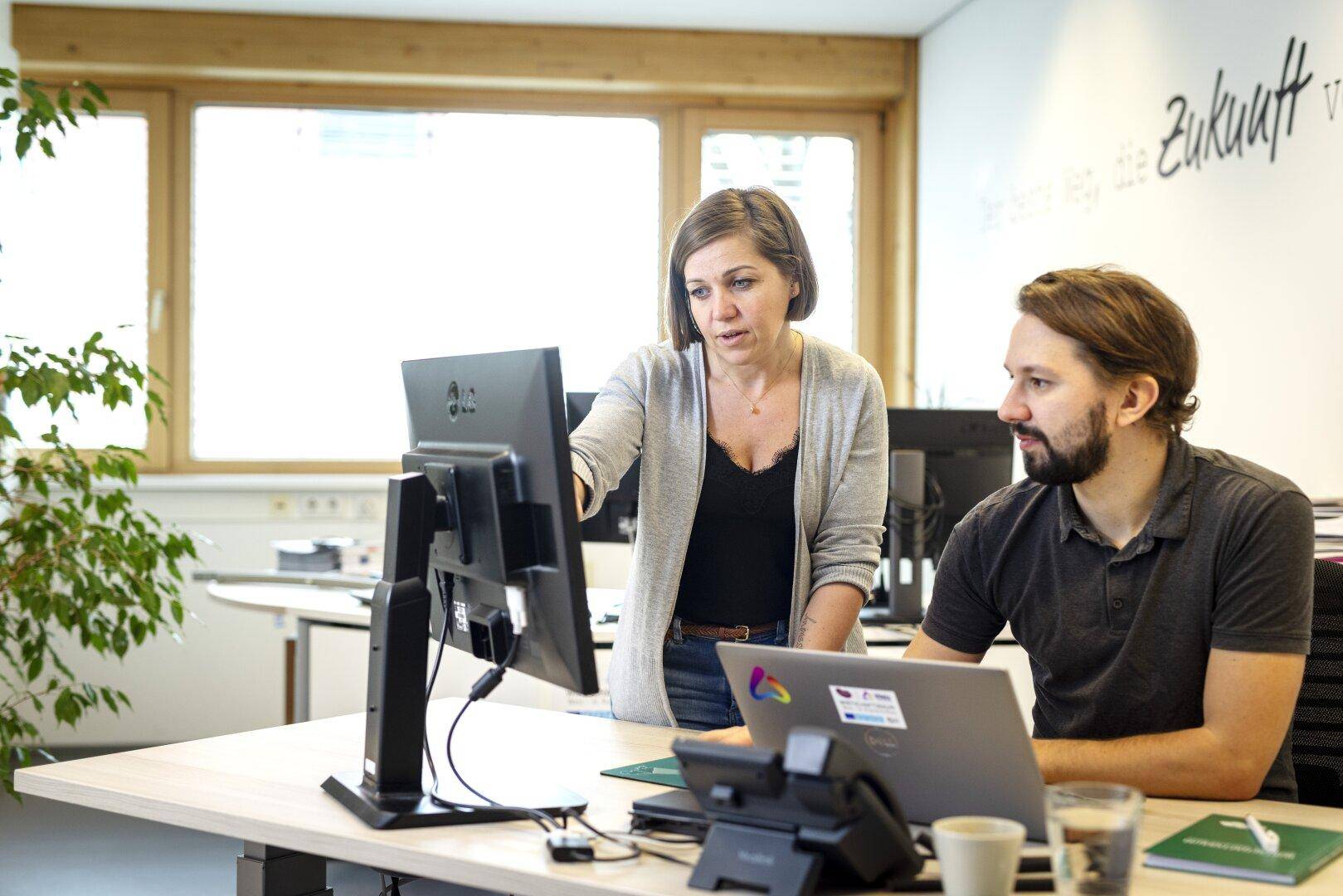 Zwei Kollegen arbeiten gemeinsam an einem Computer in einem hellen Büro mit großen Fenstern und Pflanzen.