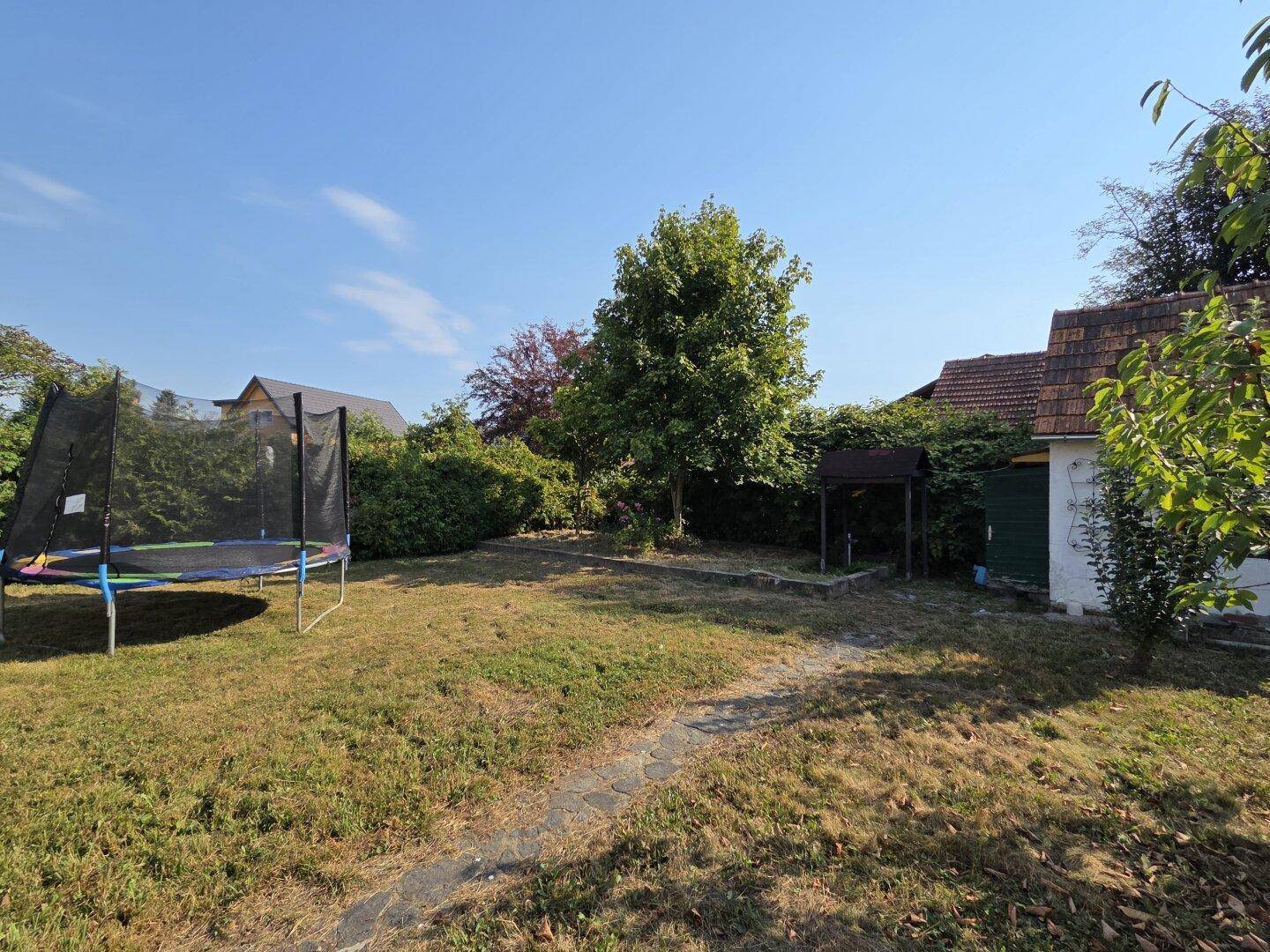 Großer Garten mit Rasenfläche, Bäumen und einem Trampolin unter blauem Himmel.