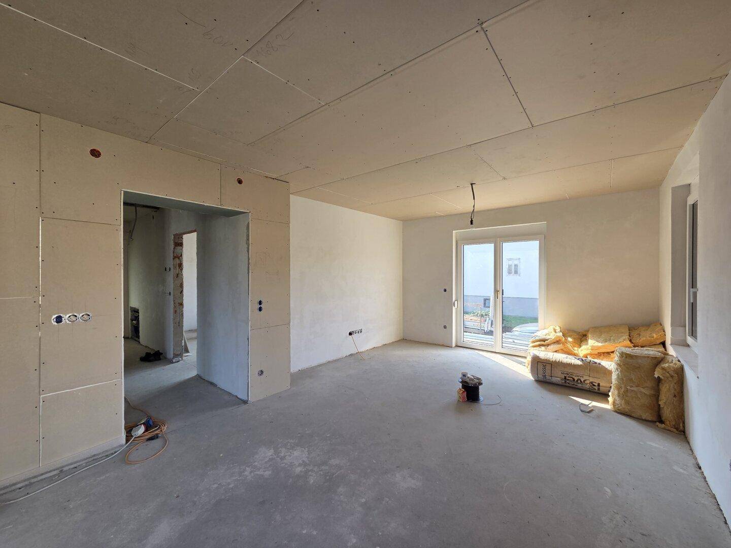 Geräumiger Wohnbereich im Rohbau mit großen Fenstern und Zugang zu einem Balkon.