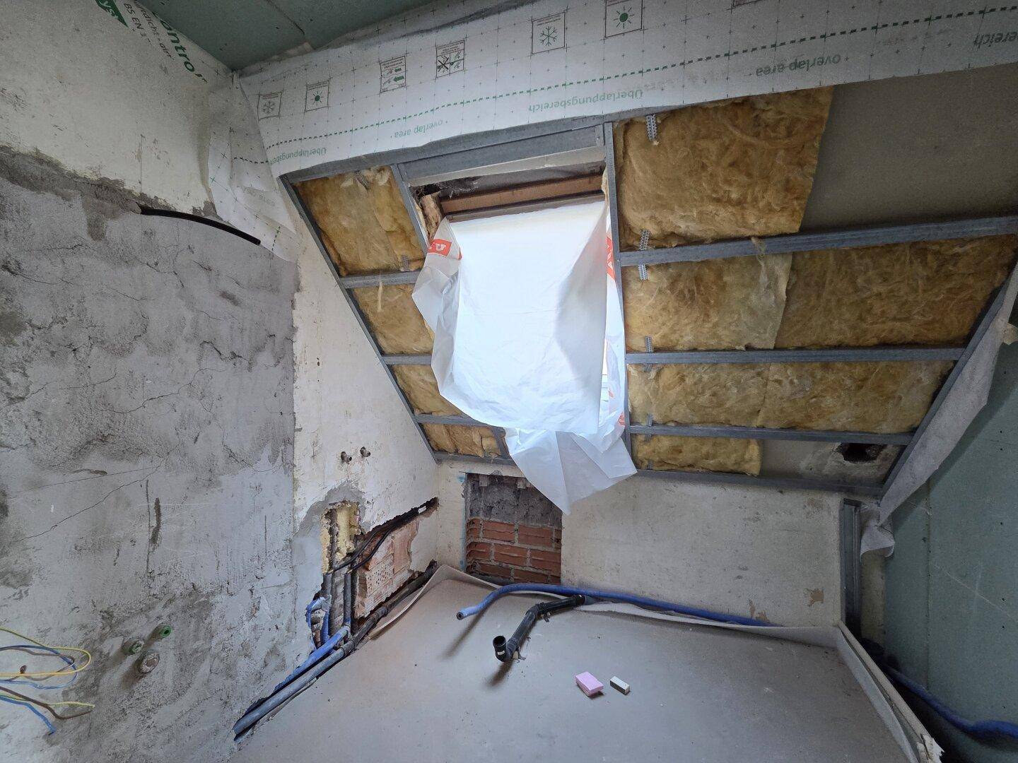 Badezimmer im Rohbau mit schrägen Wänden, sichtbarer Isolierung und Dachfenster.