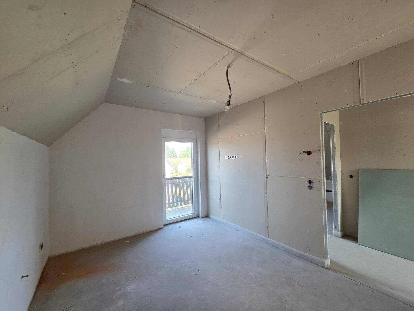 Dachgeschosszimmer im Rohbau mit schrägen Wänden und Zugang zu einem kleinen Balkon.