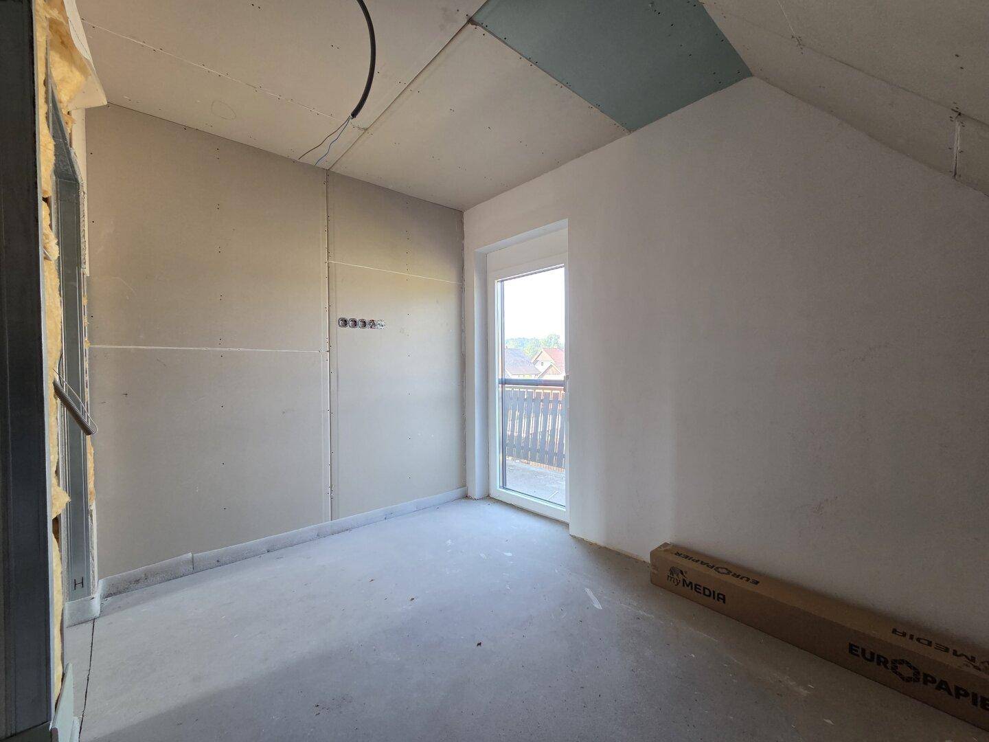 Dachgeschosszimmer im Rohbau mit schrägen Wänden und Zugang zu einem Balkon.