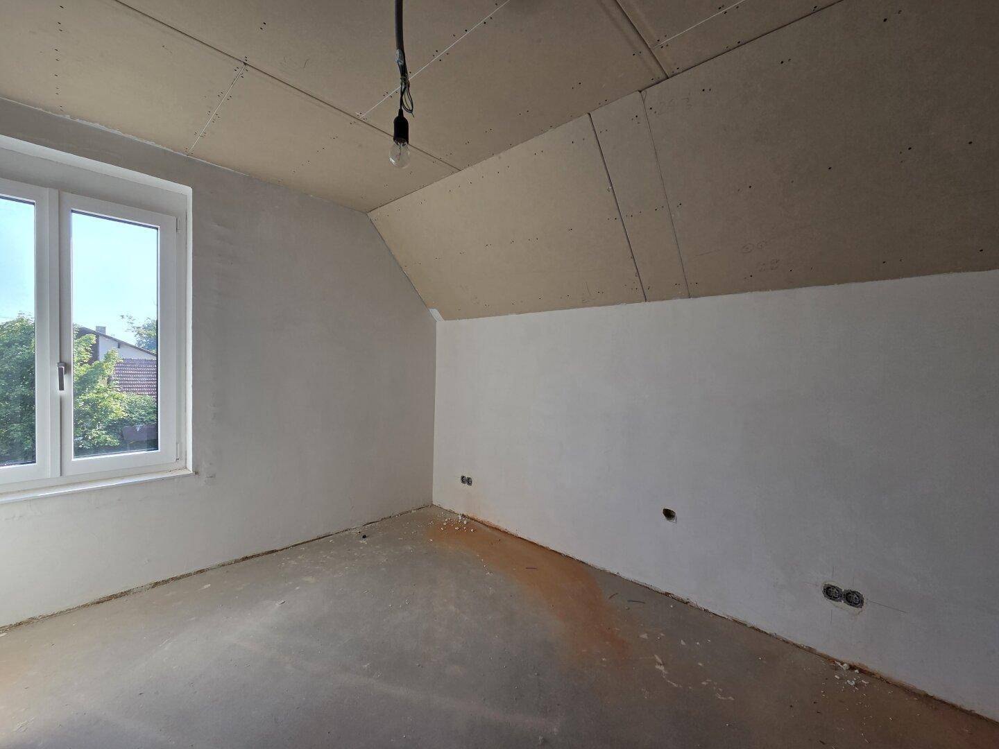 Helles Dachgeschosszimmer im Rohbau mit schrägen Wänden und Fenster.