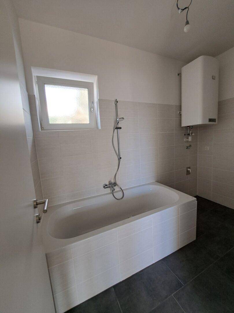 Helles Badezimmer mit Badewanne, Fenster und Wandheizkörper, ausgestattet mit modernen Fliesen.