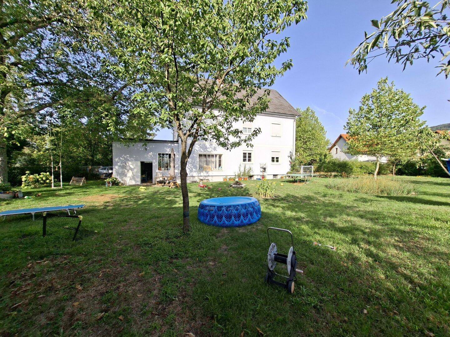 Ansicht des Hauses vom Garten aus mit einem blauen aufblasbaren Pool und viel Grünfläche.