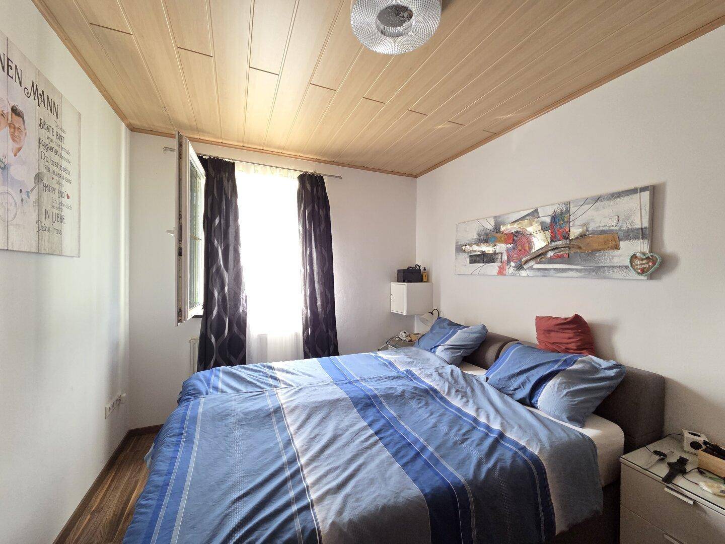 Schlafzimmer mit Doppelbett, Holzdecke und Fenster mit dunklen Vorhängen.